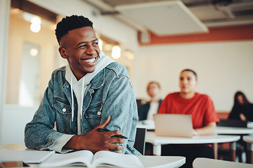 Young male student sitting in classroom holding pen in hand and books laying in front of him on desk.
