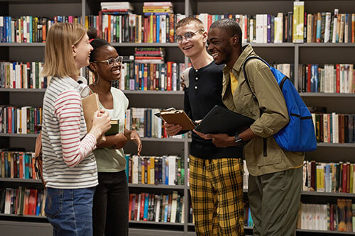 Four young students standing around in library by books on shelves.